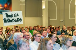 Menschenmenge sitzt im Raum, im Hintergrund ein Schild mit "Praxis tot - Patient in Not"