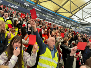 Menschenmenge auf einer Stadiontribüne mit gelben Warnwesten und roten Karten in den Händen